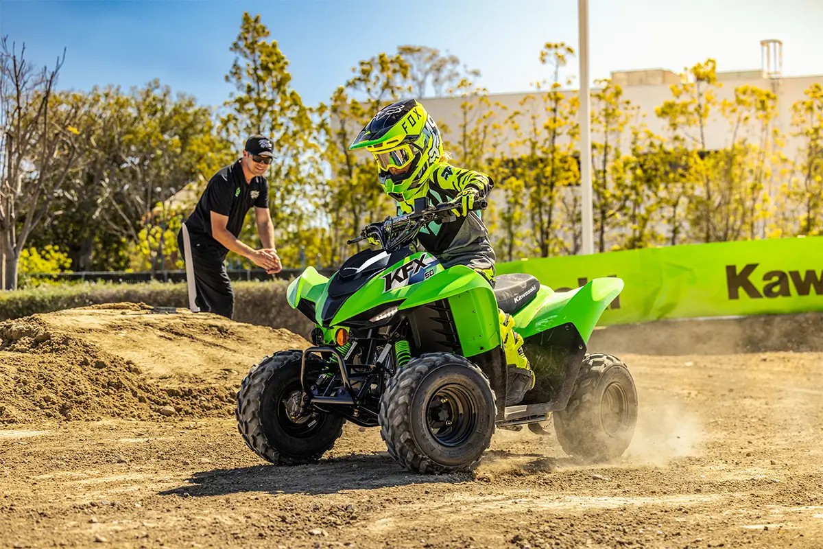 A kid riding a green youth ATV on a motocross track with a parent in the background.