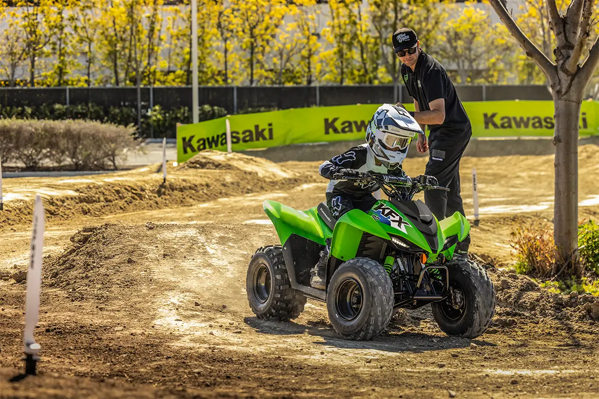 A kid riding a green youth ATV through turns on a track in front of a parent.