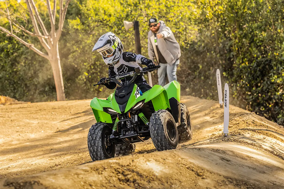 A kid riding a green youth ATV through a turn on a track as a parent gives instruction.