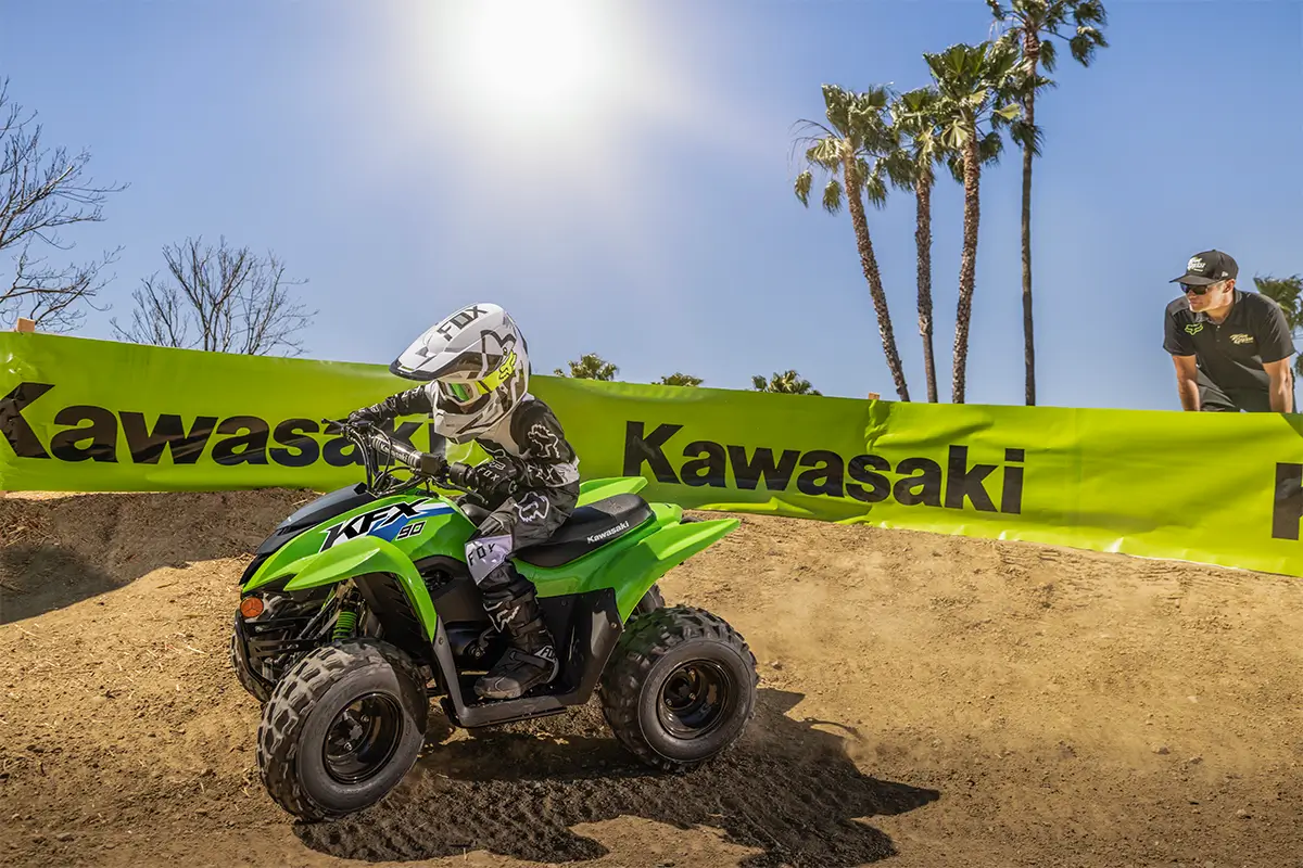 A kid riding a green youth ATV on a motocross track with a parent in the background.