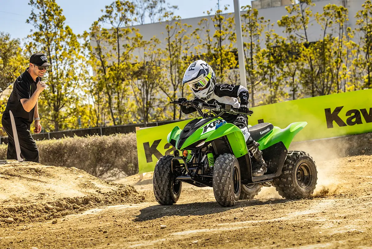 A kid riding a green youth ATV around a turn on a track with a parent in the background.