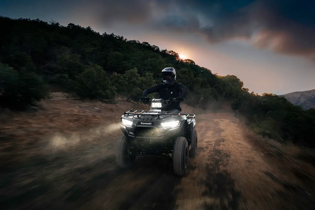 A person riding a utility ATV on a dusty road at night with headlights on.