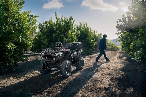 A utility ATV parked in an orchard with a person in the background walking away. opens in a new window