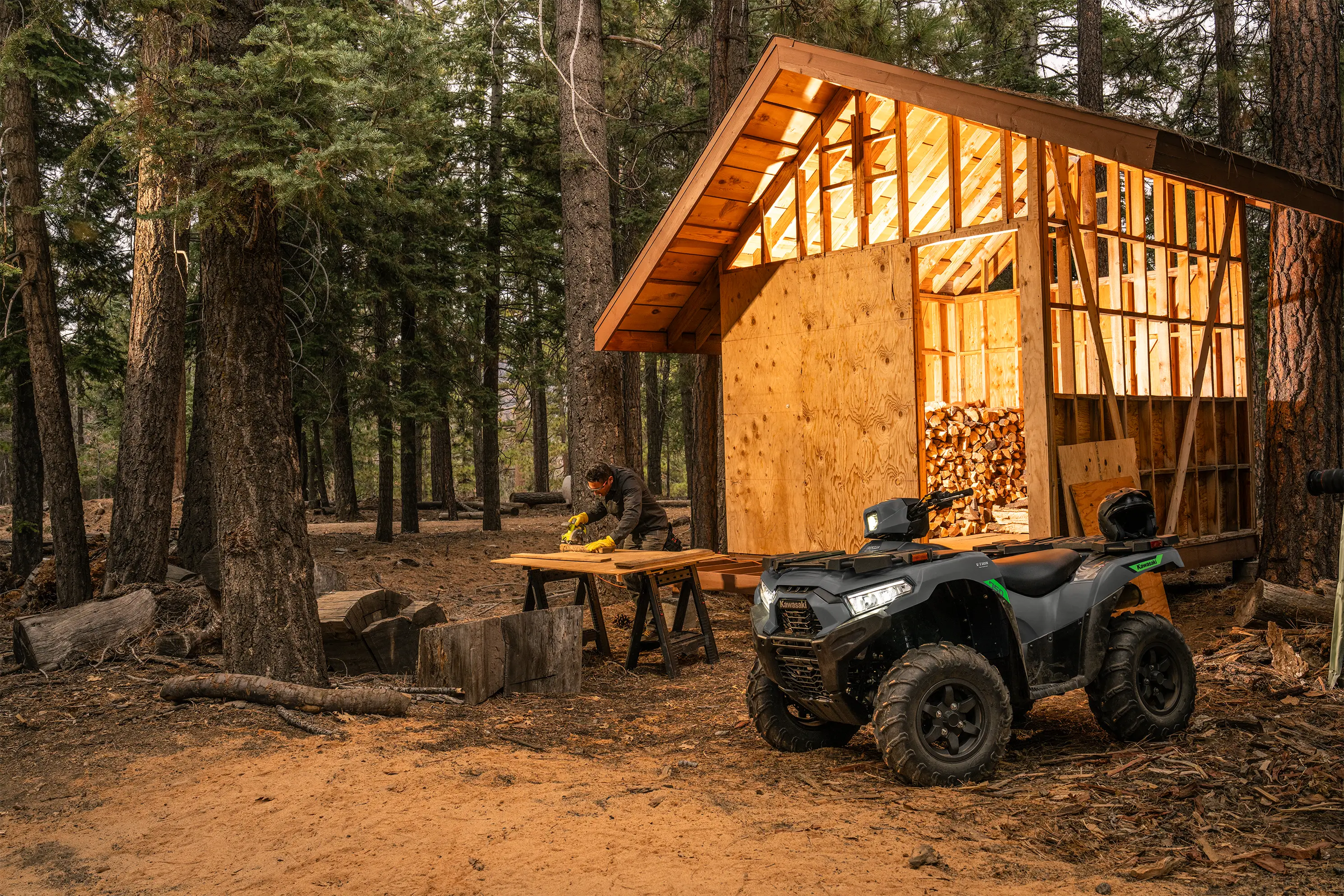 Utility ATV parked in front of a cabin under construction with a person sawing wood.