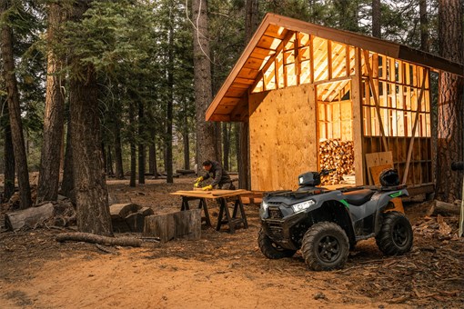 Utility ATV parked in front of a cabin under construction with a person sawing wood. opens in a new window