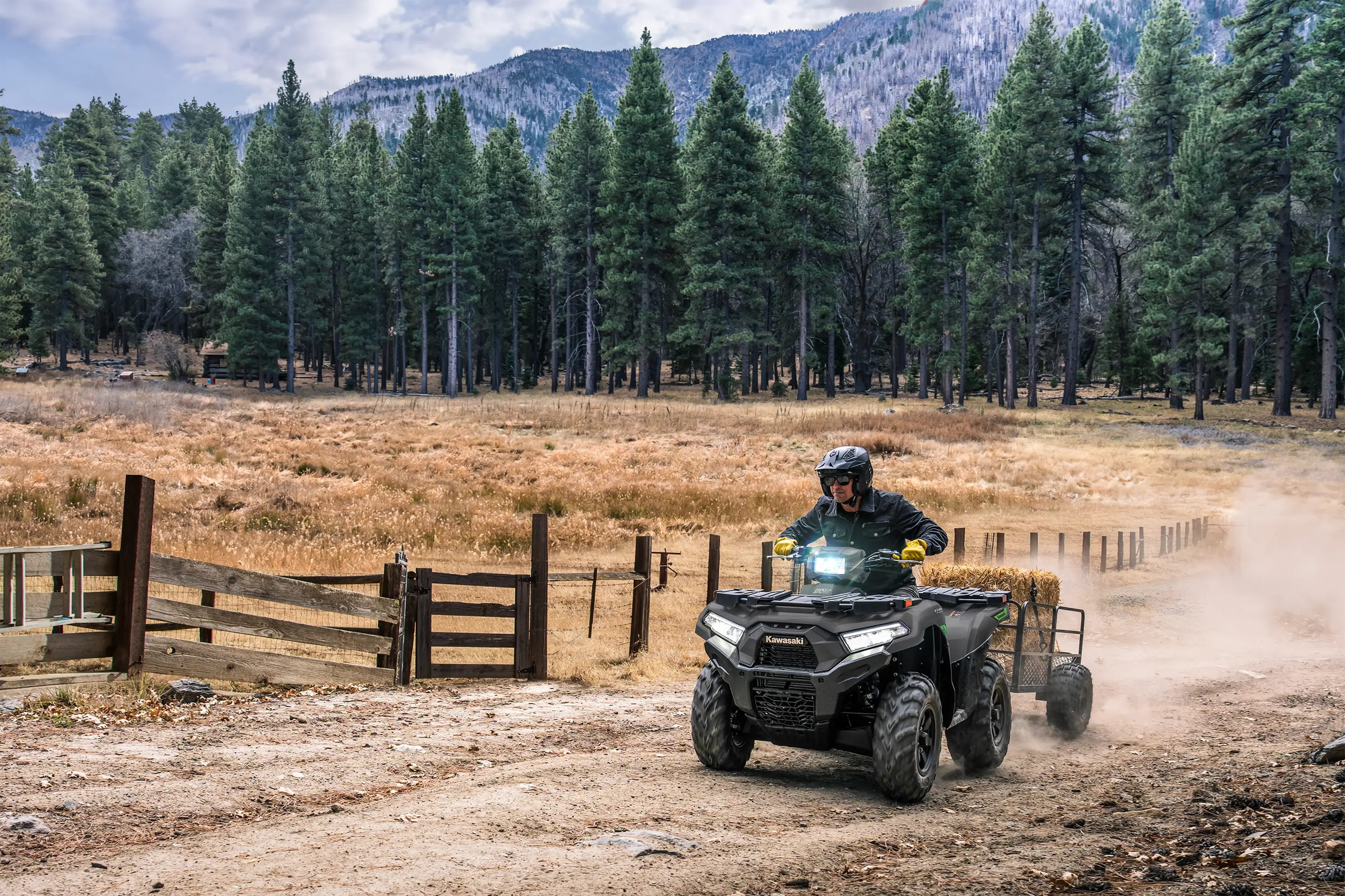 Person riding a utility ATV pulling a trailer along a fence line with a scenic forest background.