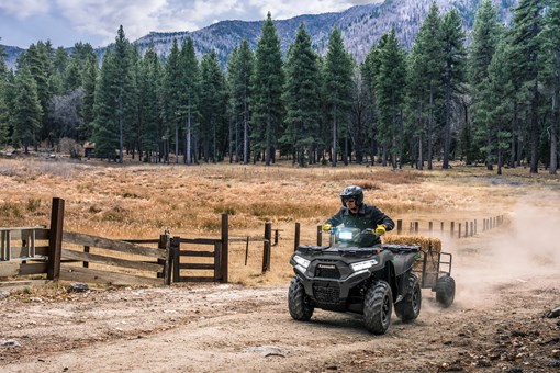 Person riding a utility ATV pulling a trailer along a fence line with a scenic forest background. opens in a new window