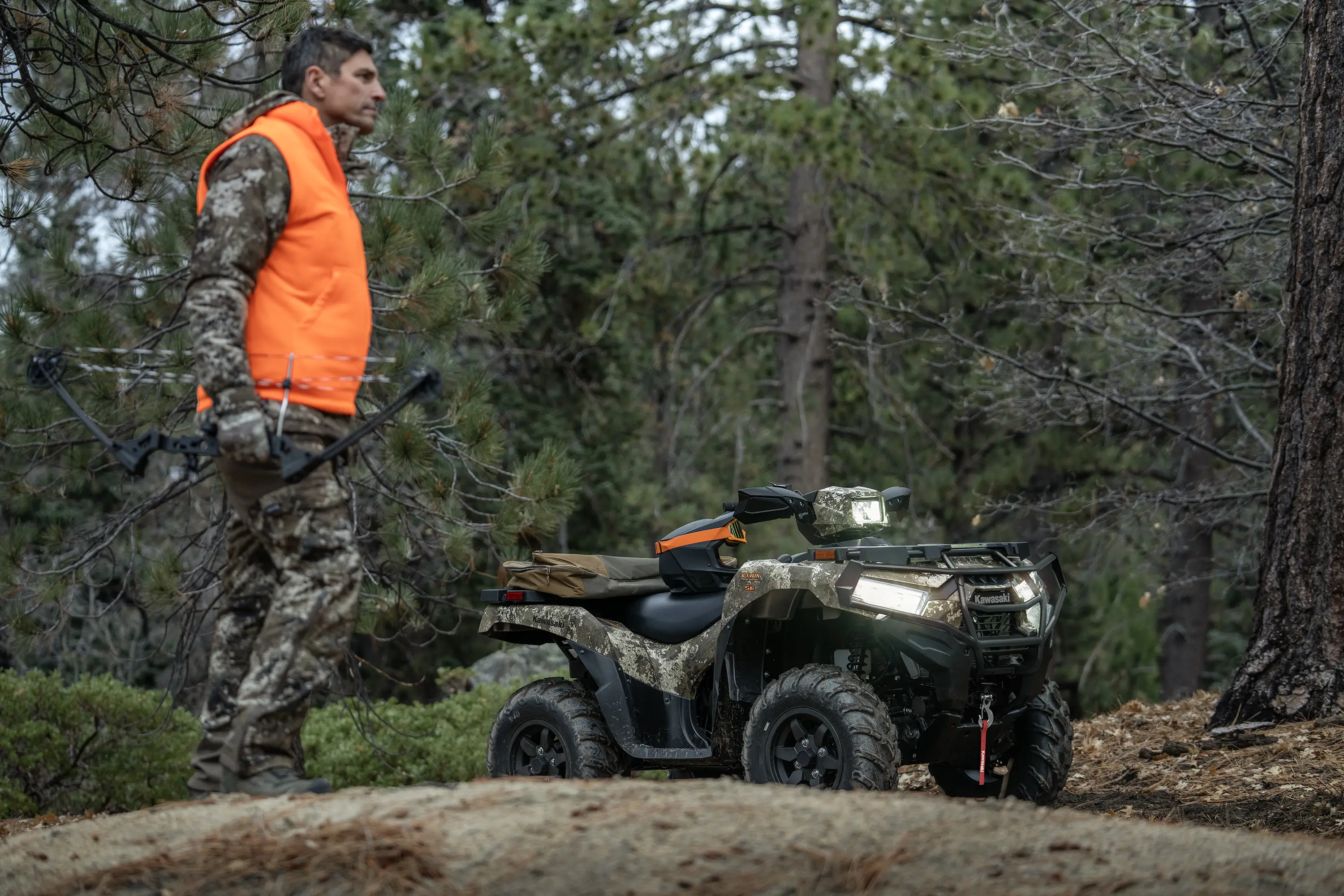 Person wearing camo and an orange vest in front of a utility ATV parked in the woods.