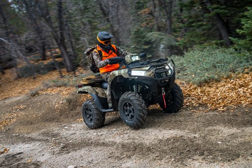 Right side view of a person wearing an orange vest riding a utility ATV on a road through the woods. opens in a new window
