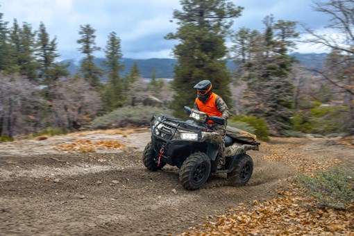 Person wearing camo and an orange vest riding a utility ATV on a road through the woods. opens in a new window