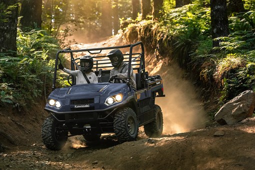 Oncoming view of two people riding in a side x side down a forest road. opens in a new window
