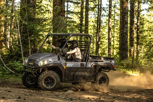 Left side view of two people riding in a side x side on a forest road. opens in a new window