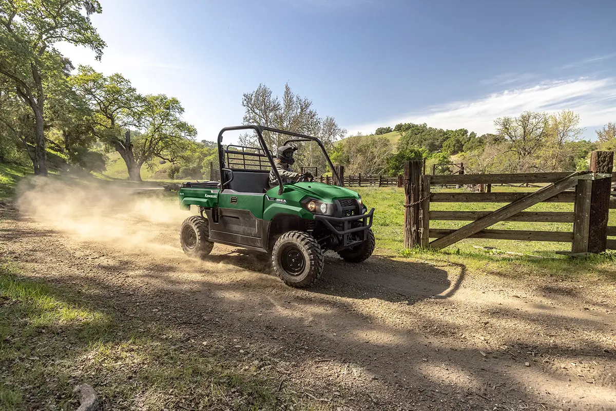 Side view of a person driving a green side x side down a dusty road.
