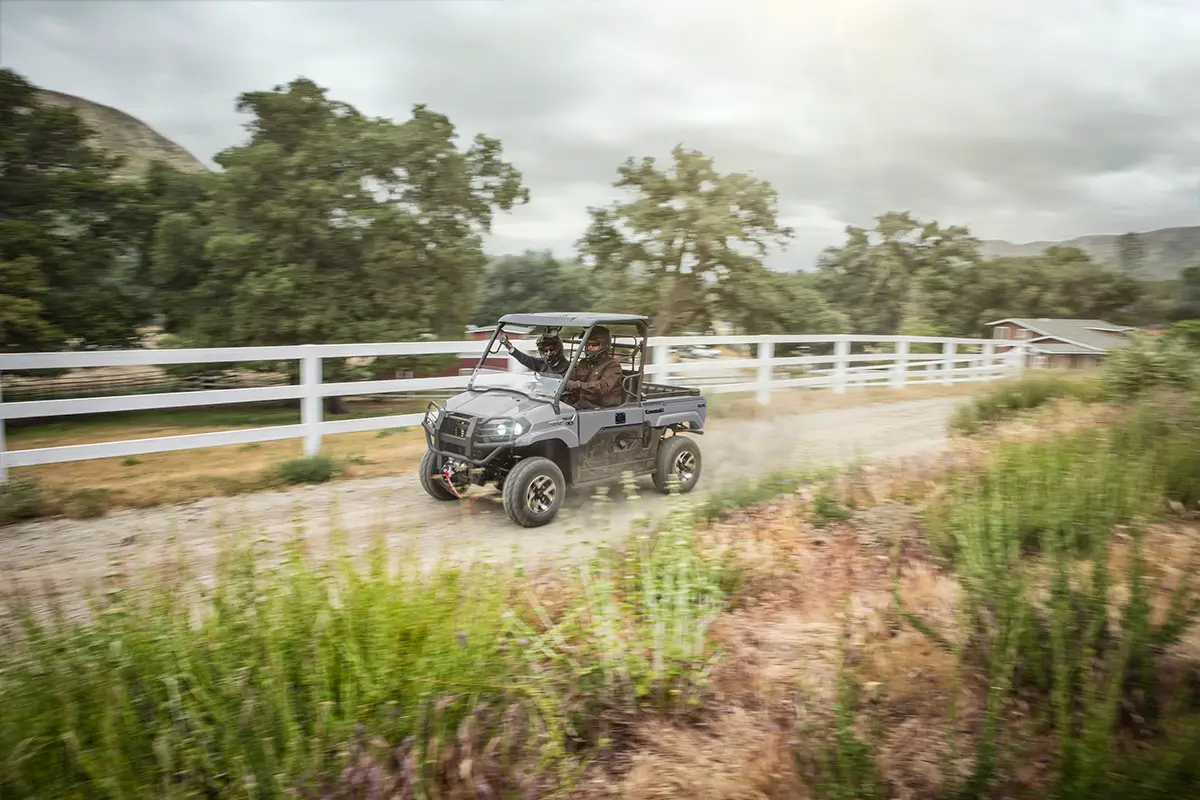 People riding in a side x side on a dirt road in front of a white fence.