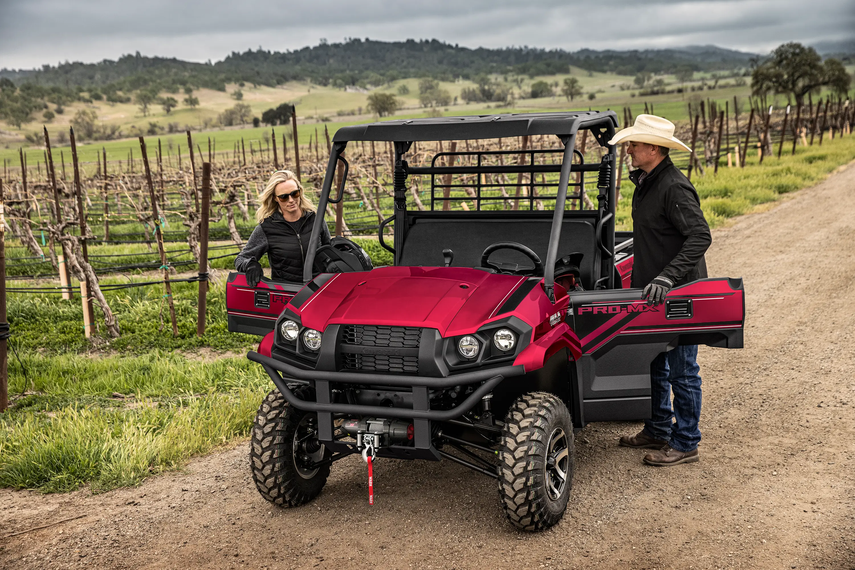 Two people preparing to get into a red side x side parked in front of grape vines.