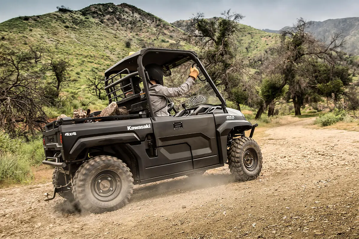 Side view of a side x side being driven down a dirt road with trees.