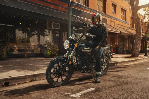 Three-quarter front angle of a person seated on a motorcycle parked in a downtown area. opens in a new window