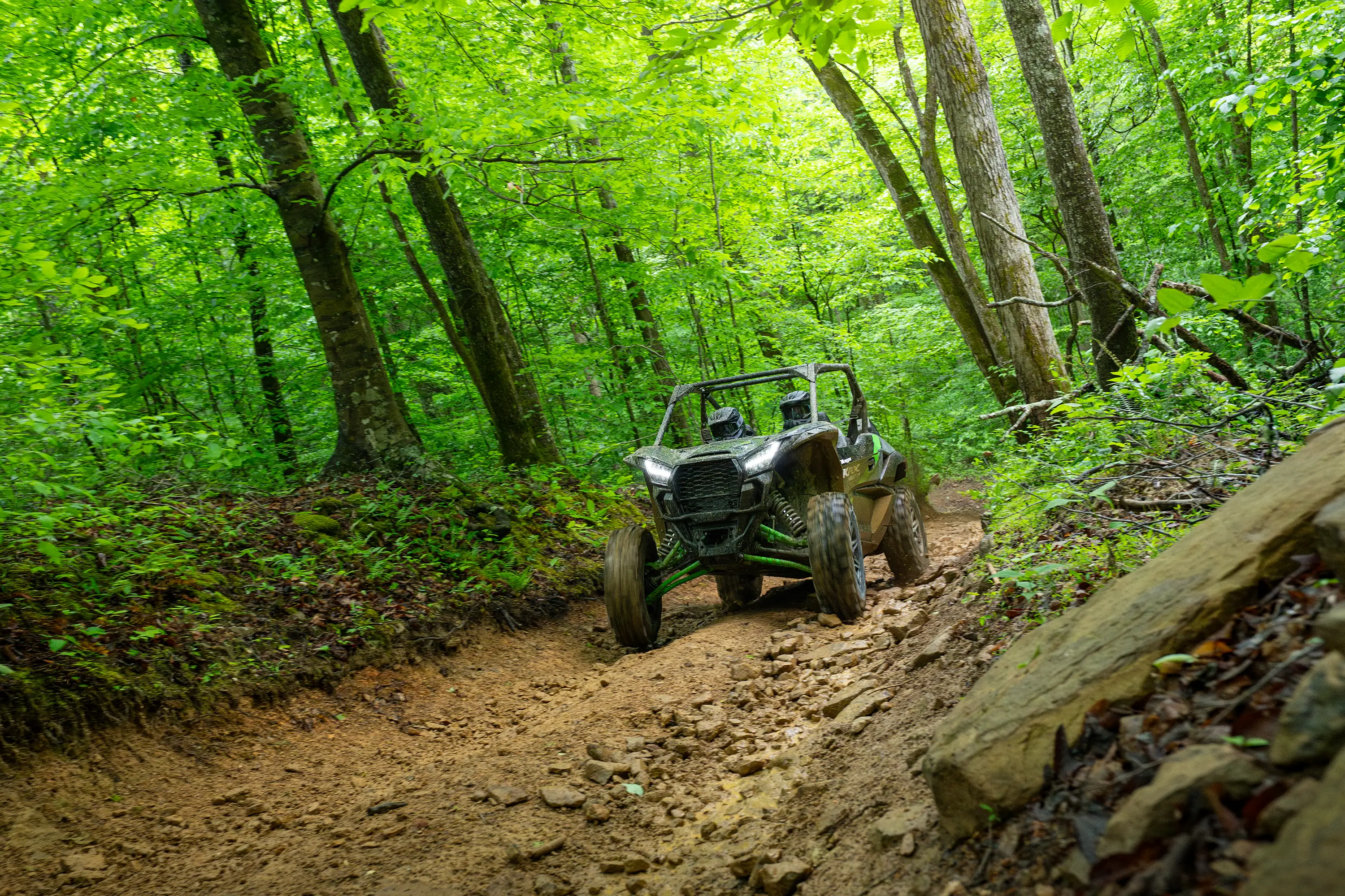 Three-quarter front angle of a driver and passenger riding a black side x side on a wooded trail.