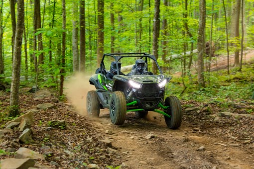 Three-quarter front angle of a driver and passenger riding a side x side on a wooded trail. opens in a new window