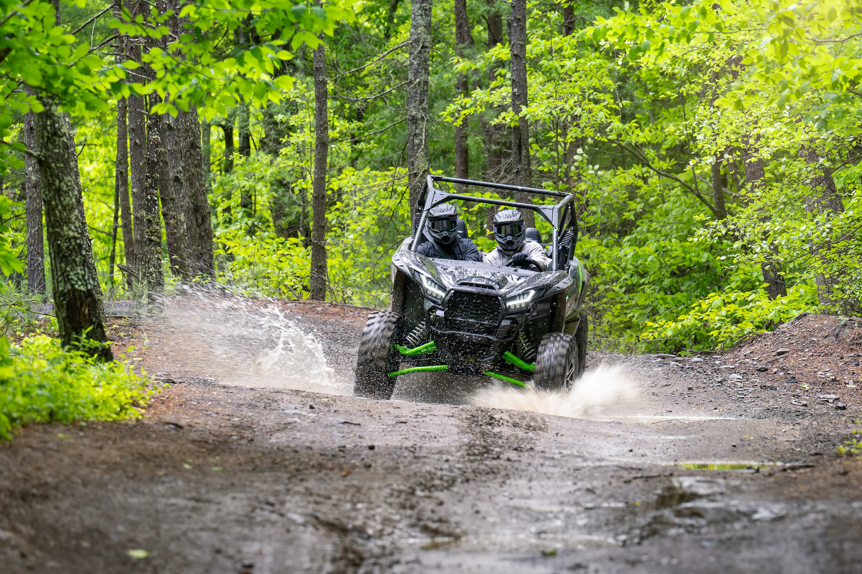 A driver and passenger riding a side x side through a puddle on a wooded trail.