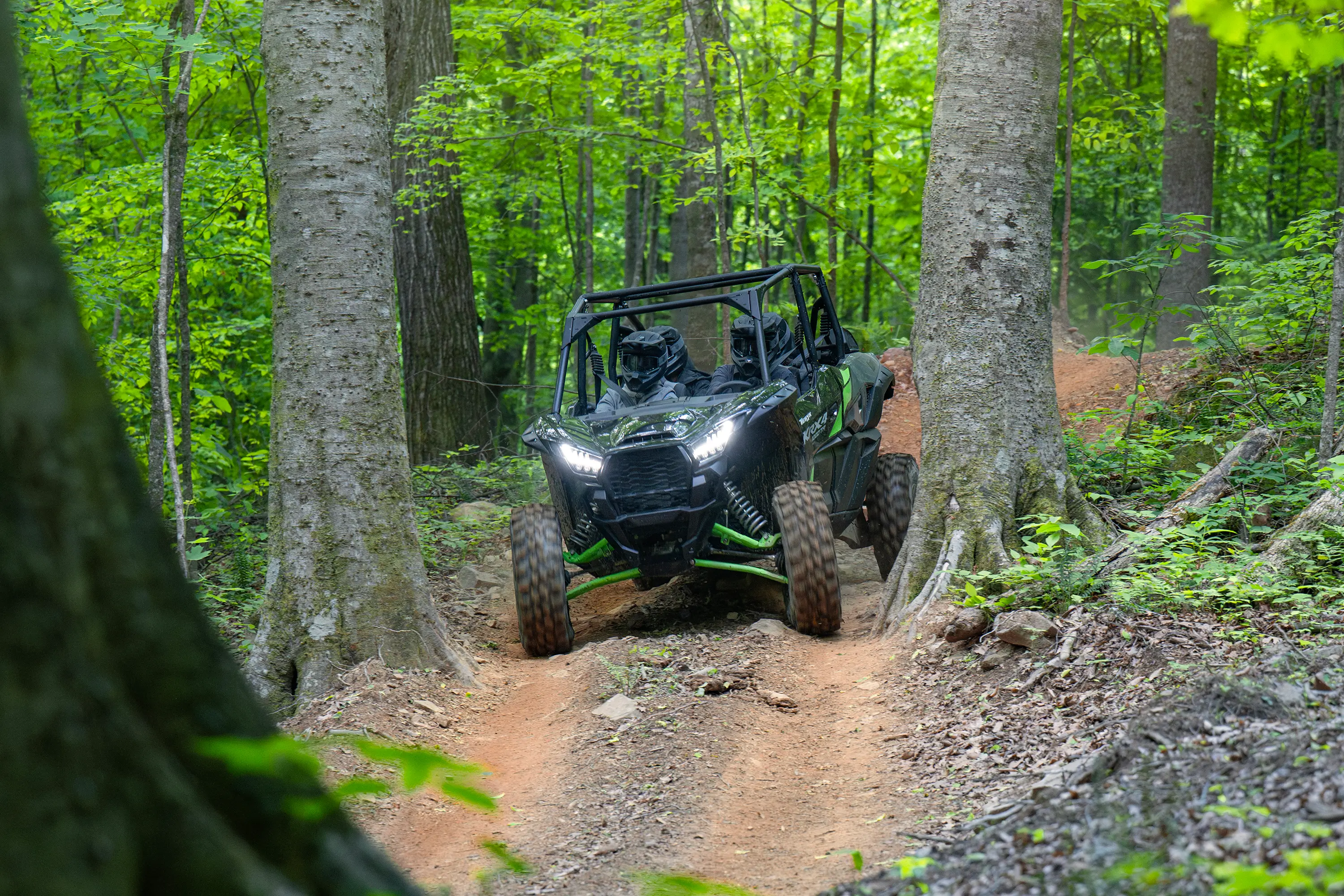 Front angle of a driver and passengers in a side x side riding down a hill on a wooded trail.