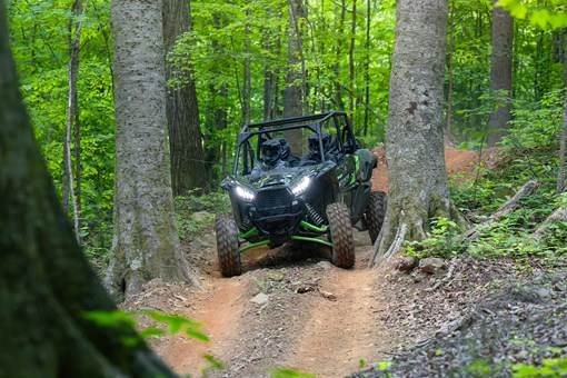 Front angle of a driver and passengers in a side x side riding down a hill on a wooded trail. opens in a new window