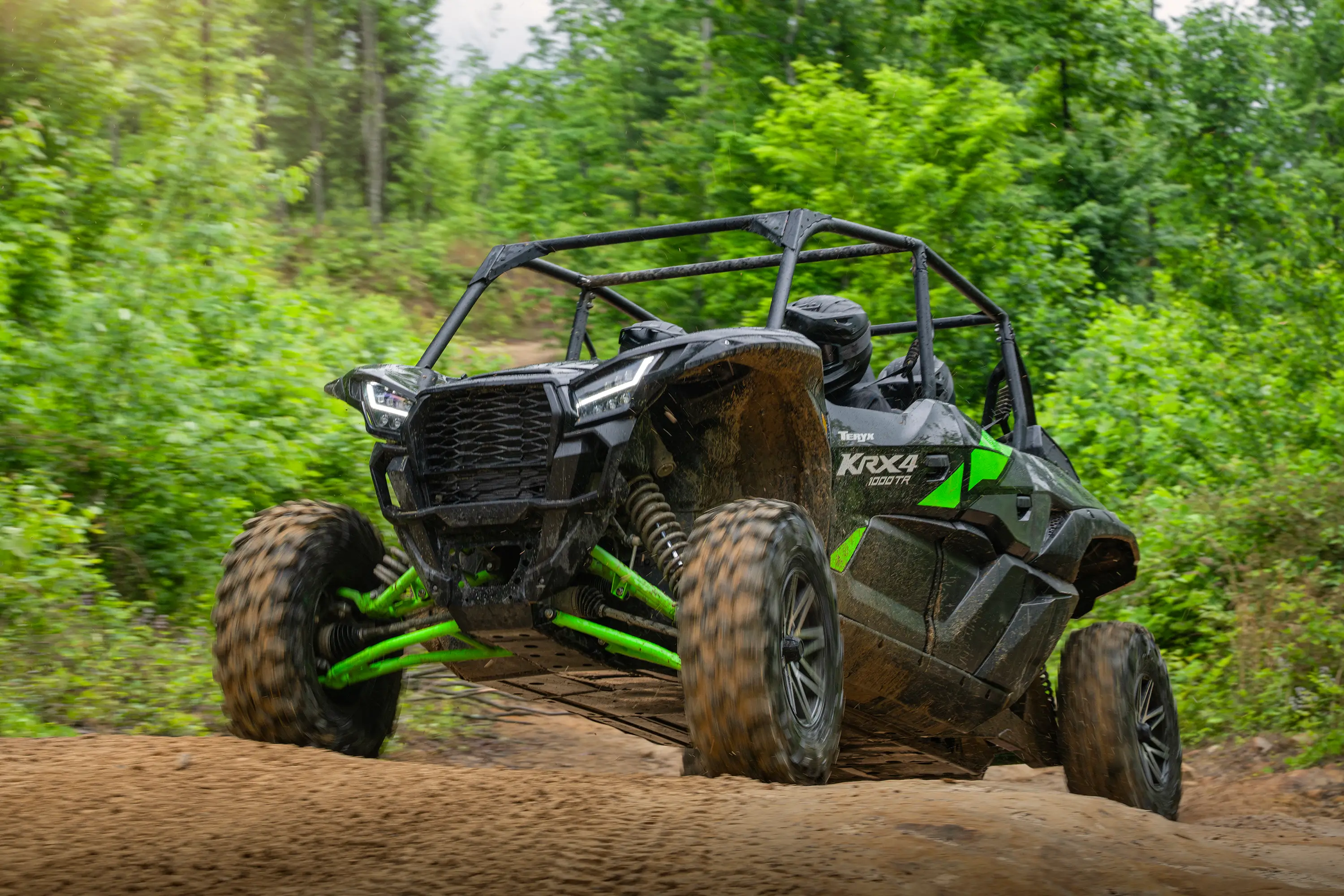 Three-quarter front angle of a driver and passengers in a side x side scaling a small hill on a wooded trail.